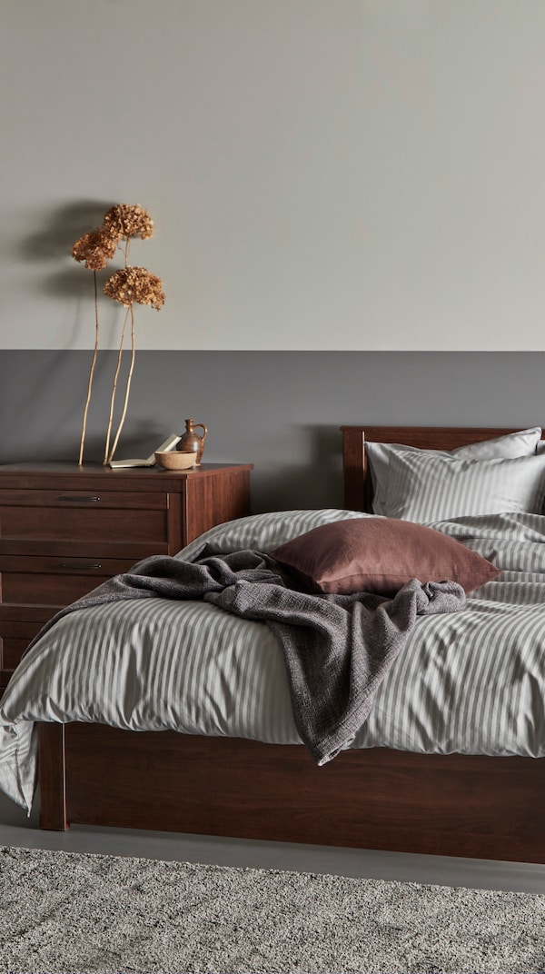SONGESAND棕色橡木床架与灰色的床单在卧室地毯在地板上和SONGESAND旁边有抽屉的柜子前与其他装饰物品。