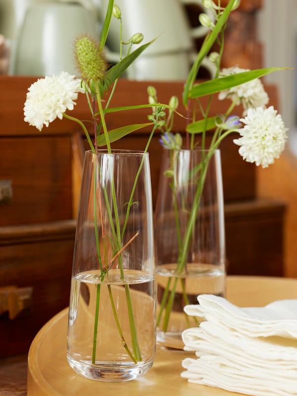两个BERAKNA透明玻璃花瓶与白色的花在桌子上用白色折叠ANSELINA餐巾纸。