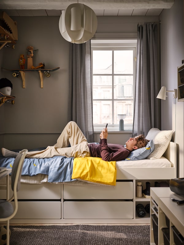 一个年轻人躺在SLAKT床上存储的窗口,阅读设备。货架由滑板在墙上。