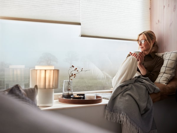 一个女人坐在一个广泛的窗台上,看起来朦胧的一天,而崎岖的毯子,靠垫和一杯茶。