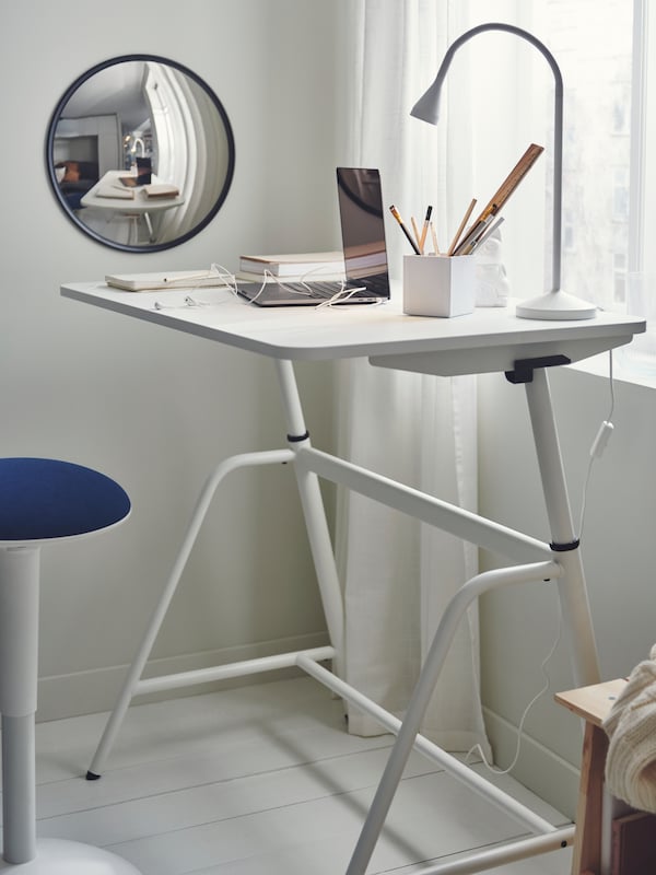 白色GLADHOJDEN坐/站桌子放在房间的角落里,笔记本和笔,和墙上的一面镜子。