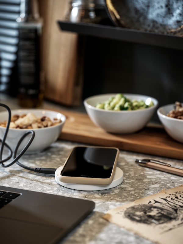 手机躺在白色LIVBOJ无线充电器的笔记本电脑之间的厨房工作台面和碗的食物。