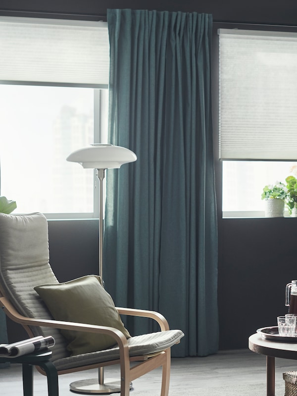 一个POANG扶手椅坐在客厅的两扇窗户PRAKTLYSING蜂窝无线百叶窗和窗帘。