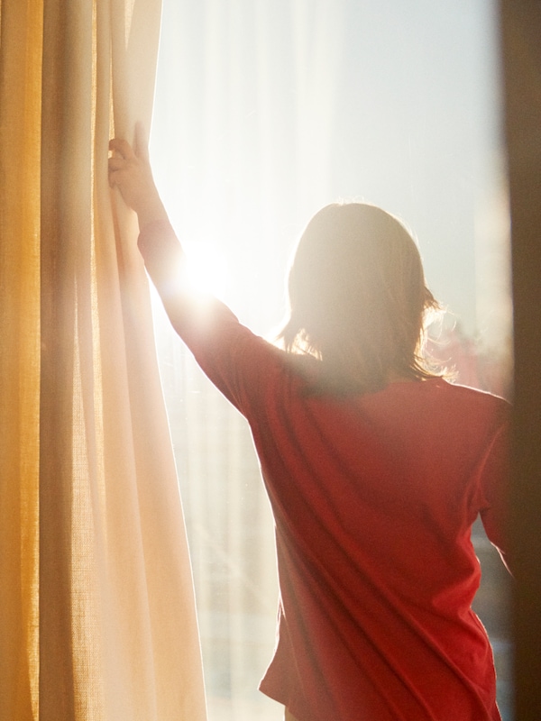 Kind in rood t恤trekt gel -met-witte gordijnen open, zodat het zonlicht naar binnen stroomt。