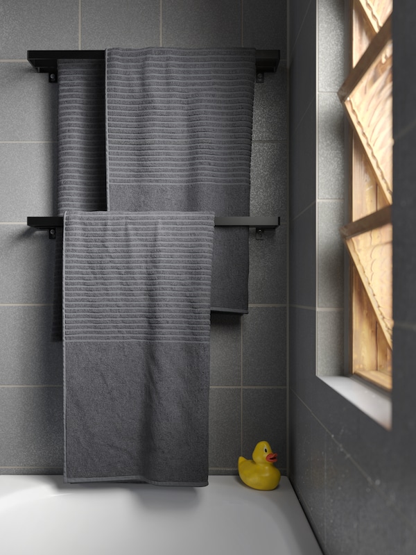 灰色的毛巾挂在两个固定在墙上的黑色毛巾rails在浴缸里。一个橡皮鸭站在浴缸里的角落。
