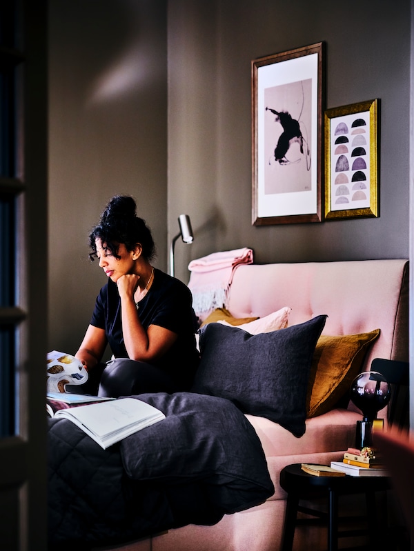 女人在卧室坐在IDANAS软垫粉红色床架与深蓝色DYTAG床单被子和枕套。旁边一个VIRRMO床头灯是安装在墙上的床架。