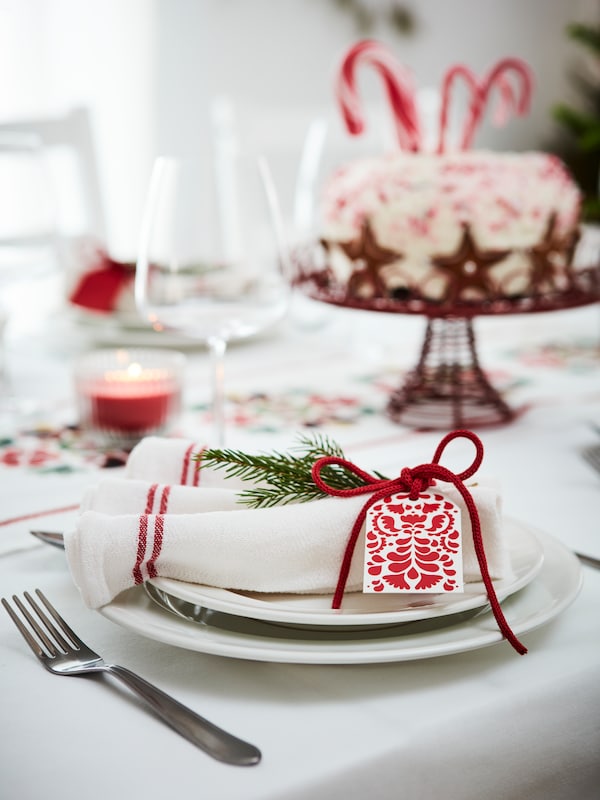 Närbild på en ljus dukning med en tygservett på tallriken och i bakgrunden syns en julfin tårta med polkagriskäppar som dekation。