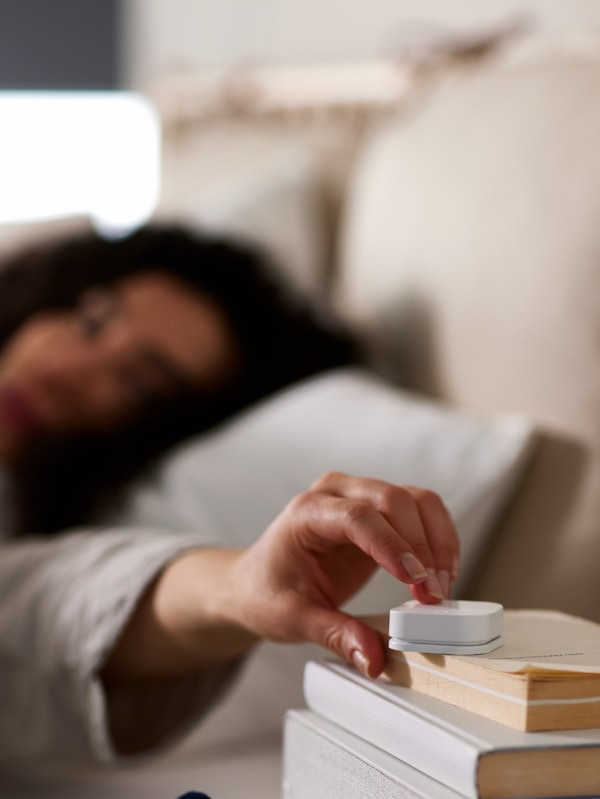 一个女人躺在床上,伸出她的手按TRADFRI无线调光器上的一些书放在床头柜上。