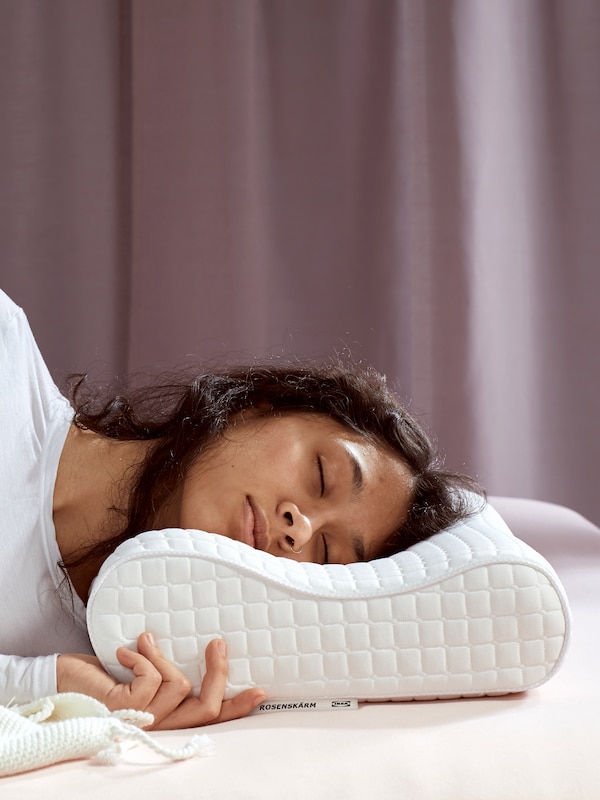 一个人穿着一件白色上衣躺睡在ROSENSKARM人体工程学的枕头在床上淡紫色的窗帘后面。