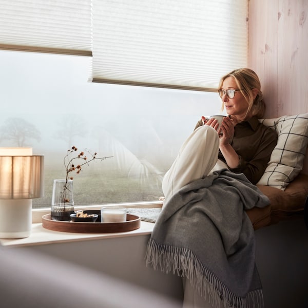 一个女人坐在一个广泛的窗台上,看起来朦胧的一天,而崎岖的毯子,靠垫和一杯茶。