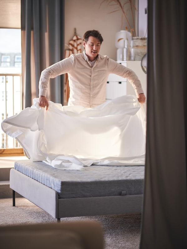 Muž pokládá bílé prostradlo DVALA na svtle edou lalounnou postel SLATTUM v loonii s velkým oknem。