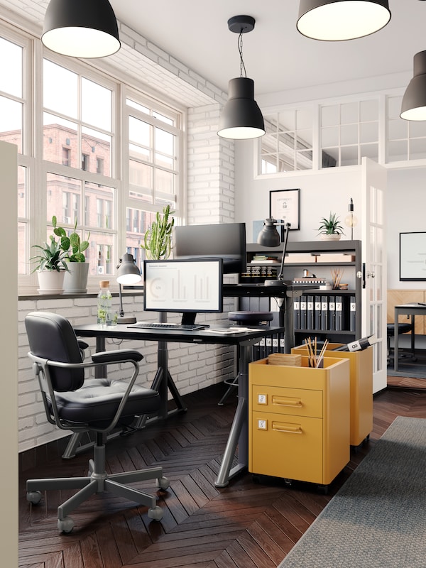 Przestronne biuro z białymi ceglanymiścianami, dwie złotobrązowe komody na kołkach IDASEN, każda obok jednego z dwoch biurek IDASEN。