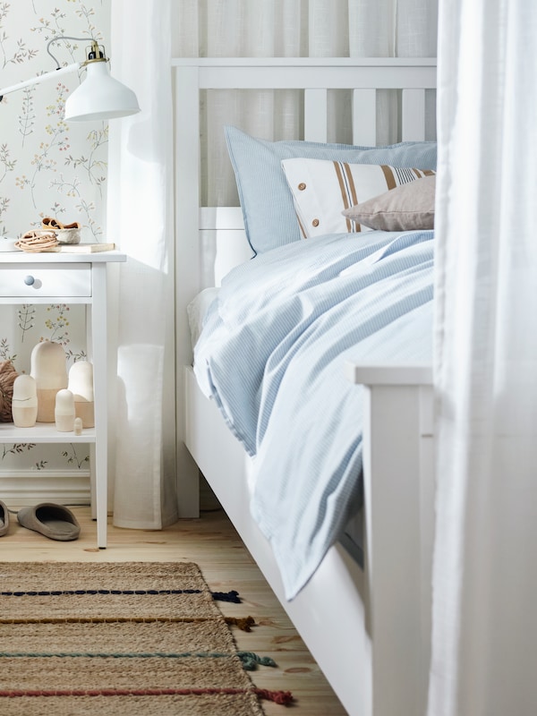 白色HEMNES床的一侧覆盖着浅蓝色的床单在一个阳光明媚的房间床头柜和一些瓶子。