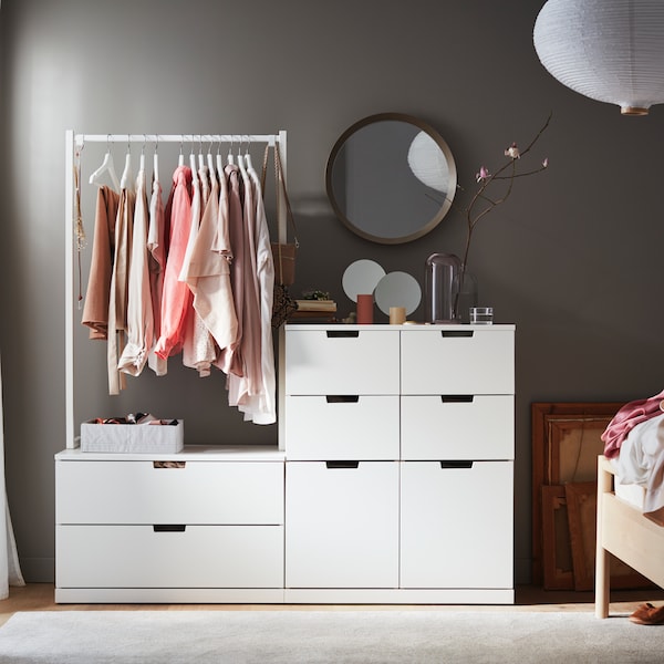 白色NORDLI有8个抽屉的柜子和一个附加挂衣杆拿衣服靠墙站在一个圆形的镜子。