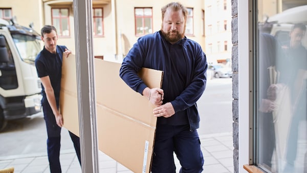 两个送货工人携带大型平面纸板盒从一辆运货卡车在街上通过一道门
