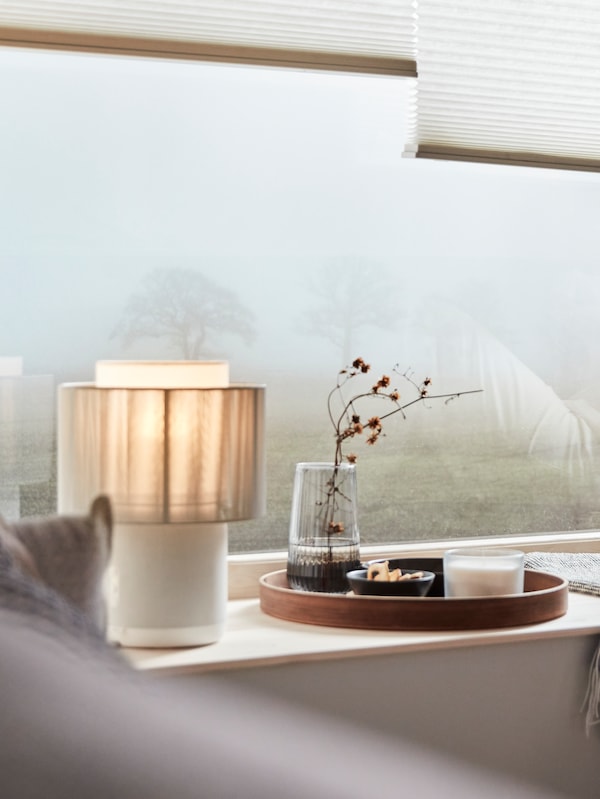 一个SYMFONISK扬声器灯与纺织品遮阳在窗台上。外面有棵树，透过雾几乎看不见。