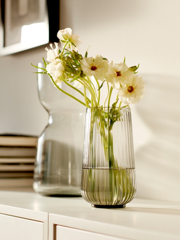 了一束白色的花在白色GRADVIS花瓶内阁,与另一个花瓶和一堆书旁边。