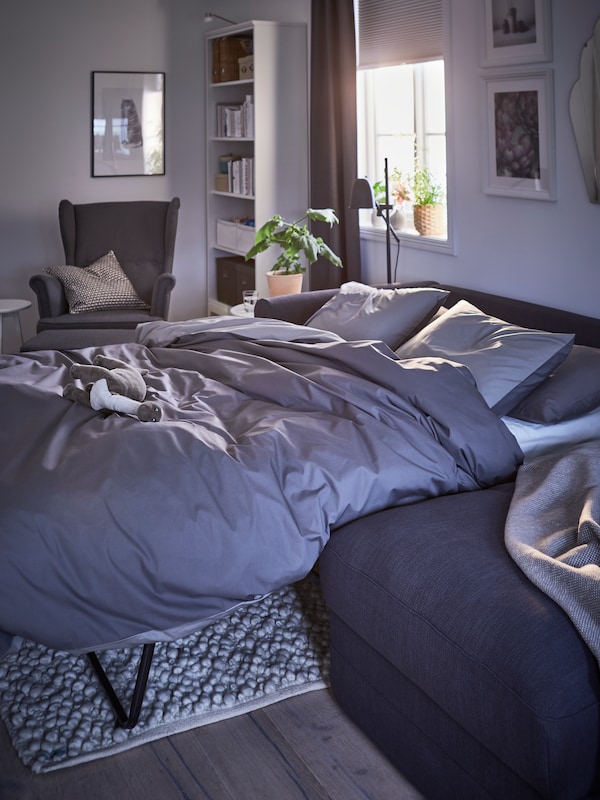 一个深灰色GRONLID 3三种座位与躺椅沙发床由灰色的床上用品的床在一个房间里,半开的盲人。
