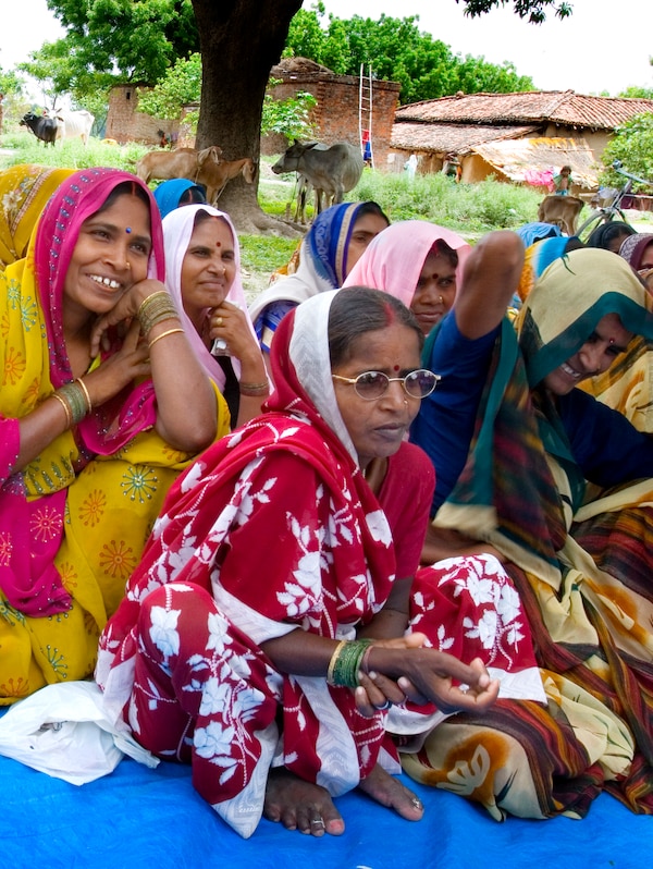 一群印度妇女在色彩鲜艳的衣服坐在外面在地毯上。在后台是一些棚屋和牛。
