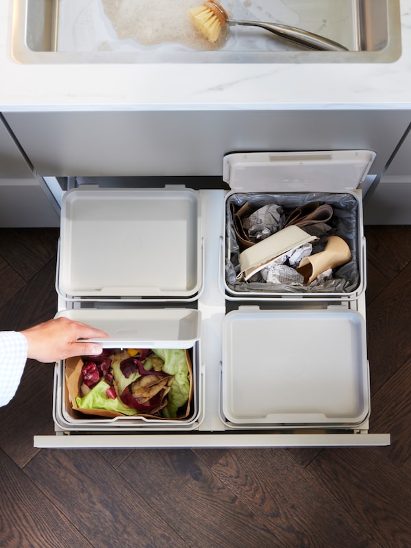 Otevřená kuchyňská zásuvka, ve které jsou vtyzki svtle šedé odpadkové kose HÅLLBAR。鲁卡otevírá jeden z kostkds potravinovým odpadem。