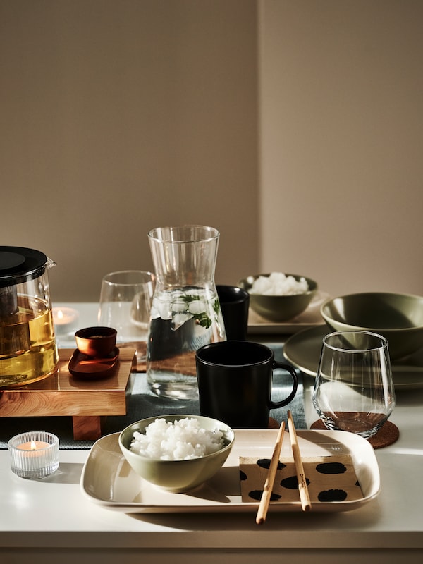 一套白色的餐桌与光滑的米色FARGKLAR盘子,马特绿色的碗,筷子,KARAFF玻璃玻璃水瓶和眼镜。