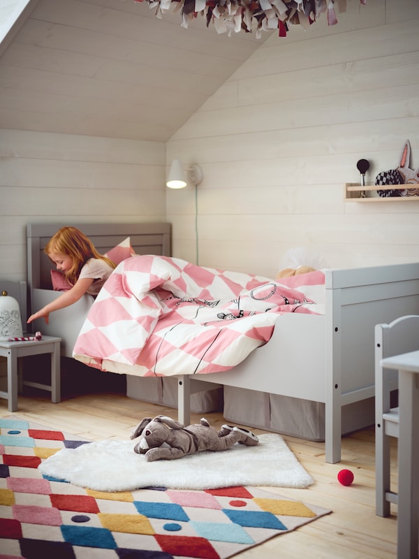 一个女孩在一个灰色SUNDVIK可扩展的床上,粉红色和白色BUSENKEL床单拿一个玩具在一把椅子在床的旁边。