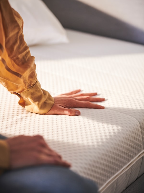 一个人坐在有ÅKREHAMN泡沫床垫的床上，用一只手往下推床垫。