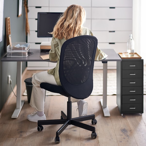 siedzizizca na czarnym krzelle biurowym FLINTAN kobieta pruje na komputerze lewitzcym na szarym biurku RODULF z regulacją wysokotzci, obok którego stoi kontenerek。