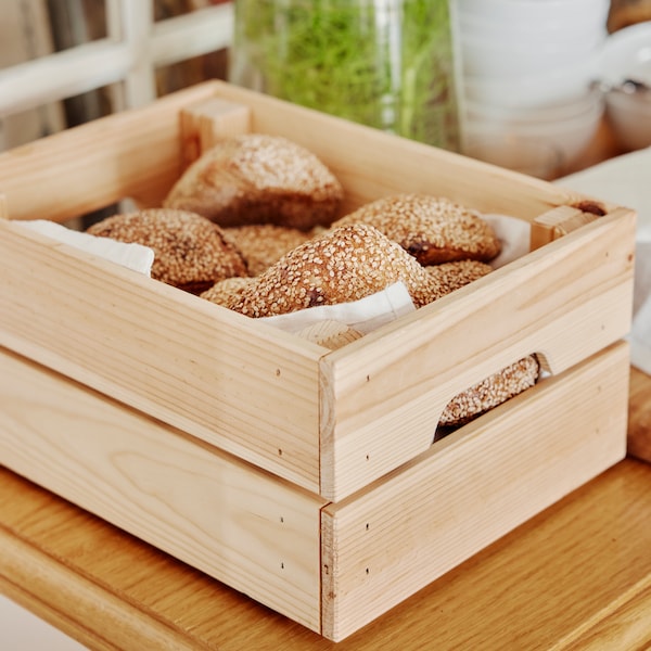 Bułki umieszczone w pudełku KNAGGLIG leżącym na drewnianym blacie, obok stoi wazon oraz ułożone w stos miseczki。