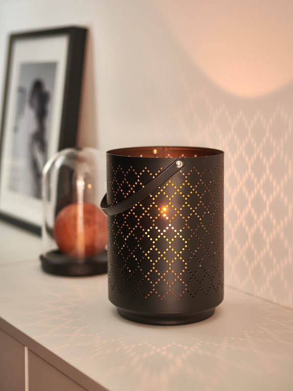 黑色URSKILJA灯笼1块内有FENOMEN块蜡烛点燃的蜡烛,旁边侧板装饰物品。