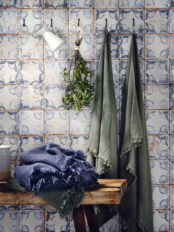 HÖGAFFEL väggkrokar på vilka ljusgröna VALLASÅN badhanddukar hänger samt en mörkblå VALLASÅN badhandduk som ligger på en träbänk。
