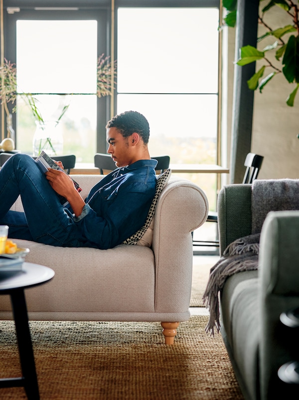明信片的人Jeanskleidung sitzt ausgestreckt auf einem hellbeigen VISKAFORS 3 er-sofa和这样Zeitschrift。