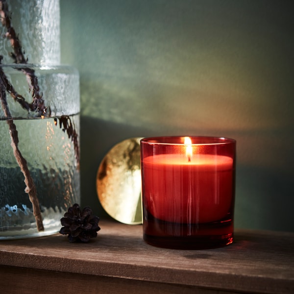 VINTERFINT kaars与de geur van sinaasappel en kruidnagel在een rood glas staat op een schoorsteenmantel naast een glazen KONSTFULL vaas相遇。