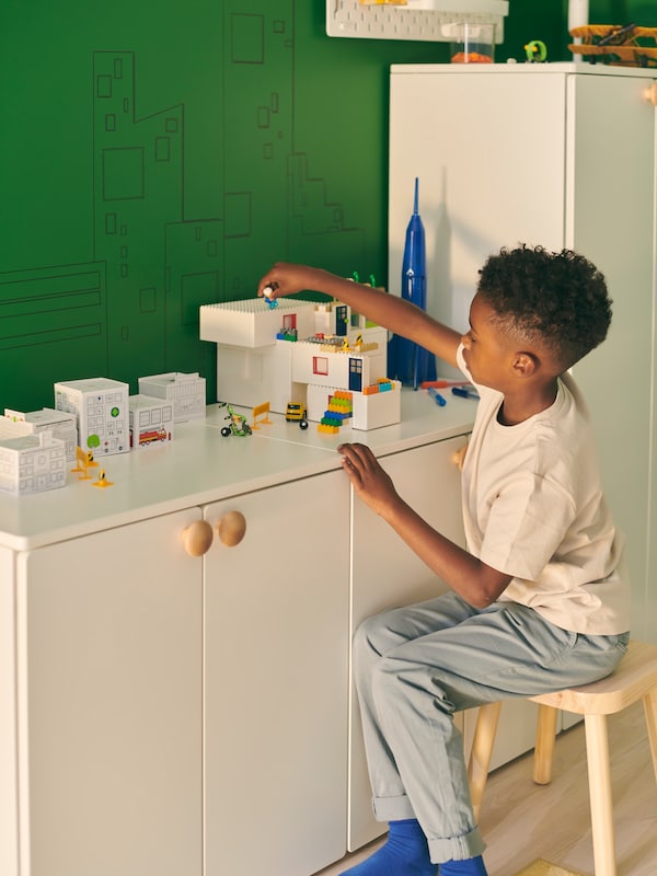 一个男孩坐在凳子上,玩一套BYGGLEK乐高砖放在白色SMASTAD cabinet-and-wardrobe组合。