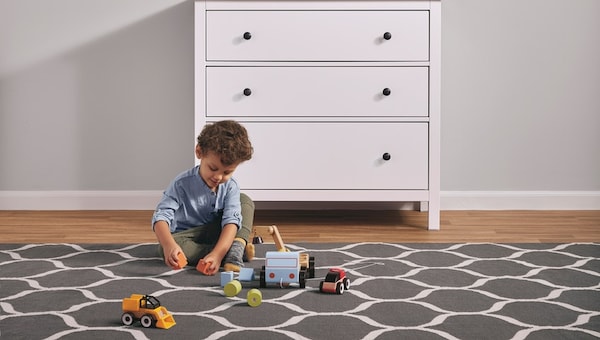 Ett litet barn som leker med leksaker på en stor, mönstrad matta bredvid en vit HEMNES byrå med tre lådor。