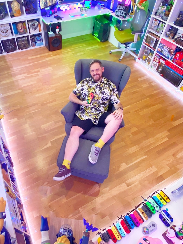一个微笑的人是靠STRANDMON扶手椅。他有一个视频游戏控制台。周围的墙壁覆盖着开放的书架上放满了游戏和玩具。
