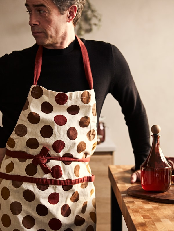 一个穿着KRÖSAMOS彩色围裙的人站在VADHOLMA黑橡木厨具旁，手里拿着KRÖSAMOS瓶子。