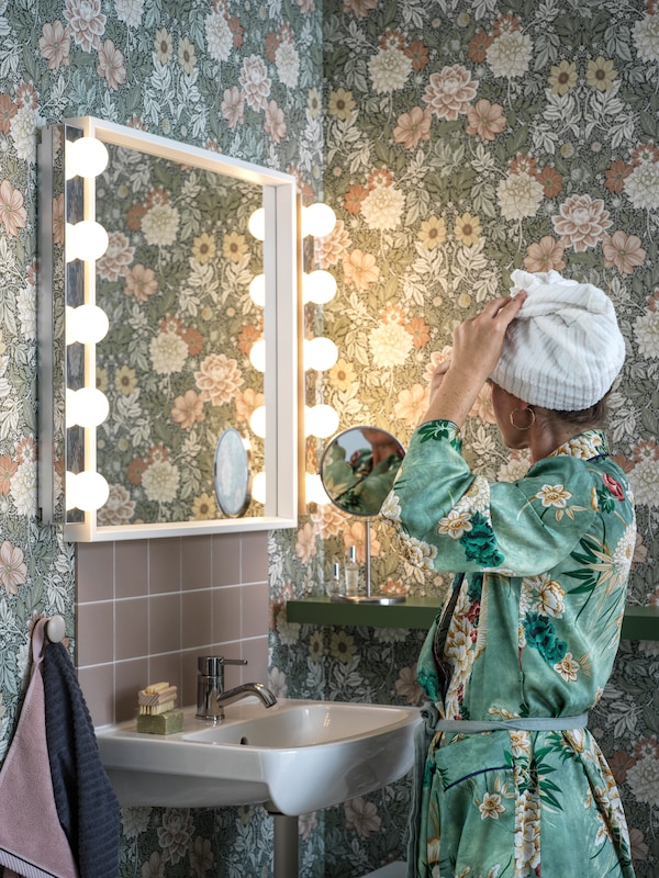 一个女人在一个长袍匹配的花卉图案浴室墙壁站在一个镜子NISSEDAL LEDSJO墙灯。