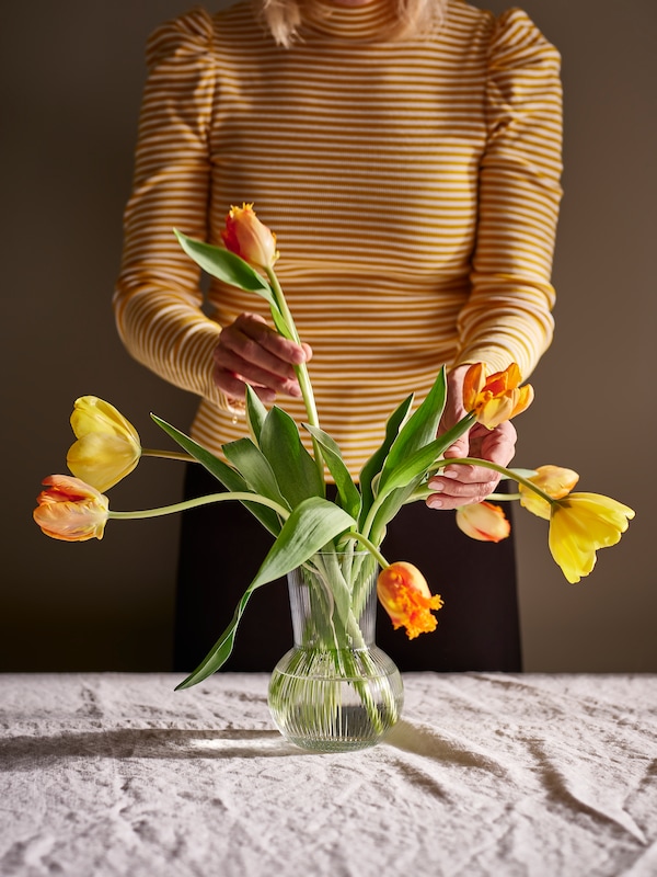 一个女人安排黄色和橙色郁金香在PADRAG清晰的玻璃花瓶natural-coloured台布的餐桌。