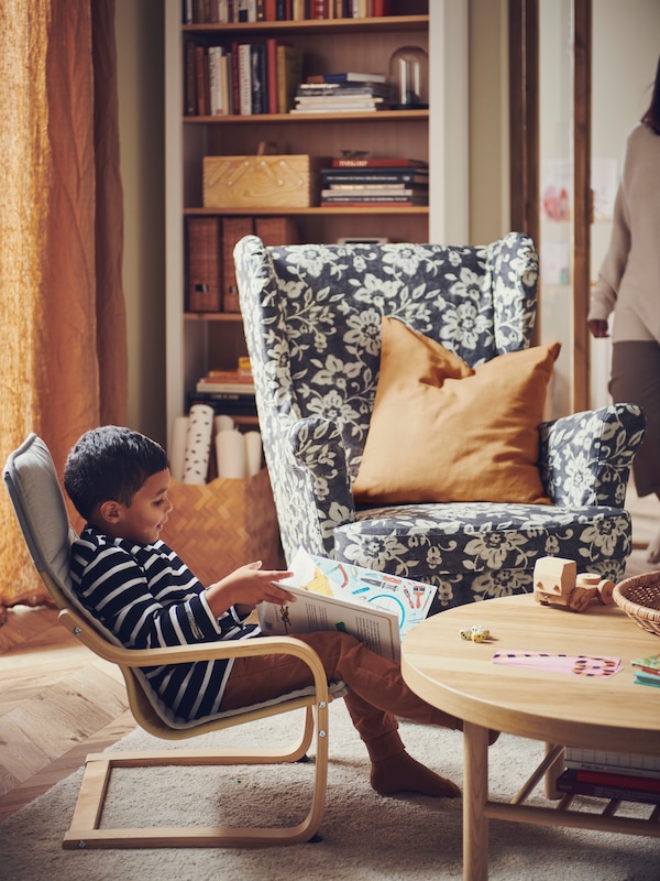 一个孩子坐在POANG儿童椅STRANDMON翼椅子旁边,一副bold-patterned客厅的沙发套。