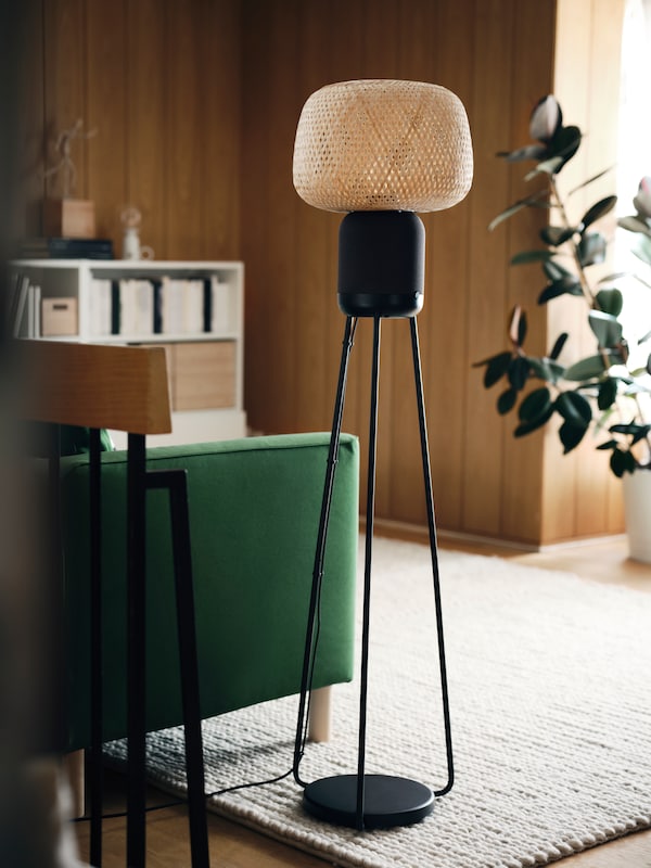 Lampa podłogowa SYMFONISK z głośnikiem wi - fi我bambusowym kloszem stoi na białym dywanie HJORTSVANG obok zielonej sofy PARUP。