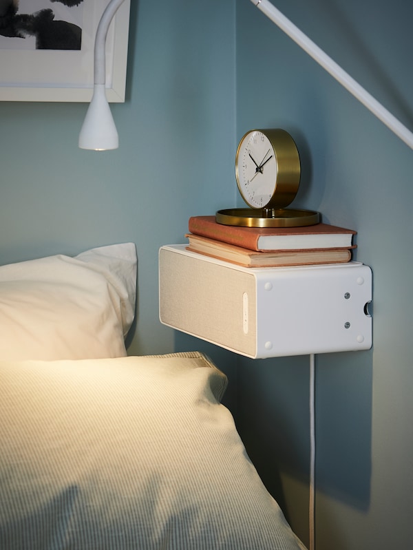 白色,SYMFONISK无线扬声器安装水平在蓝色墙壁的床上,两本书和一个金色的钟上。
