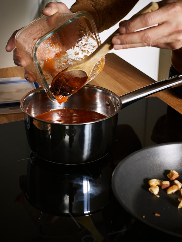 一个人的手用木匙刮番茄酱从正方形玻璃食品容器变成一个平底锅。