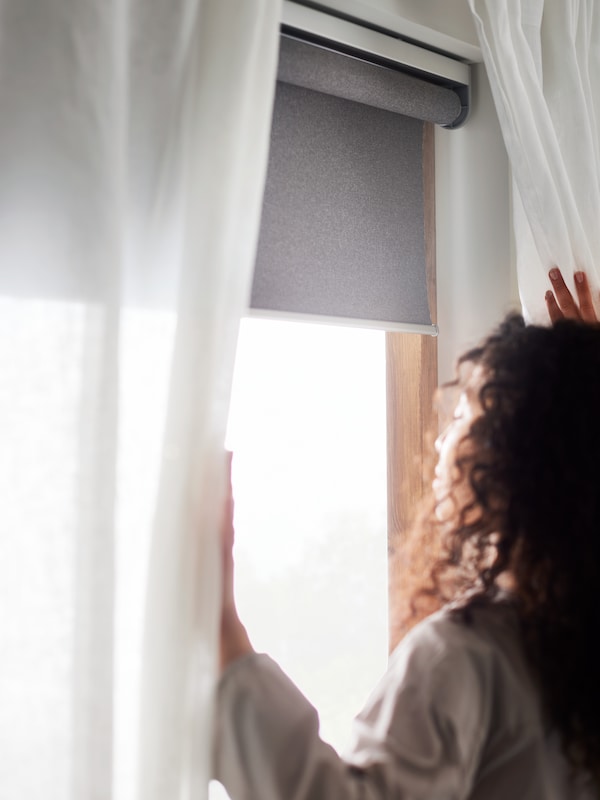 一个女人站在一个窗口与白色窗帘和一个灰色FYRTUR无线/电池屏蔽遮光窗帘。