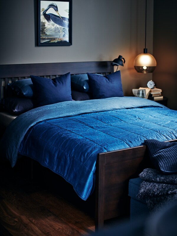 IDANAS黑棕色存储床架在黑暗的卧室亮蓝色的床单和枕头。吊灯挂在一边的床架,在罗马帝国的衣柜前昏暗的光芒。
