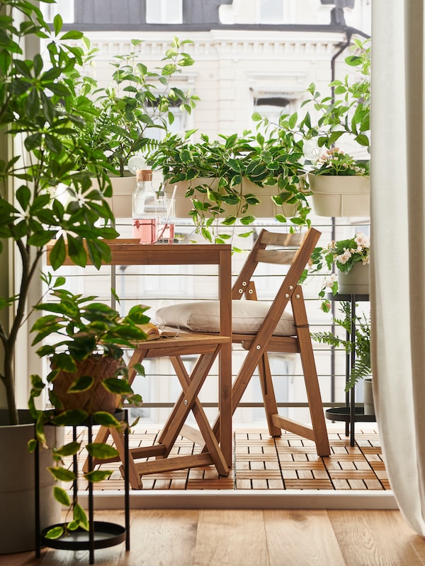 Et lyst brunbejdset NAMMARO havebord, en klapstol og sammenklappelig taburet pa en altan indhyllet我grønne potteplanter。