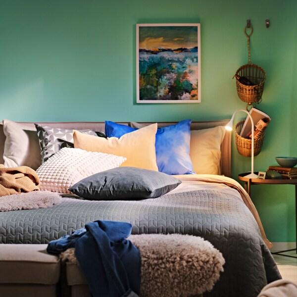 床由光grey-beige ANGSLILJA床单,床罩和各种垫子,旁边一个黑暗grey-beige托盘表。