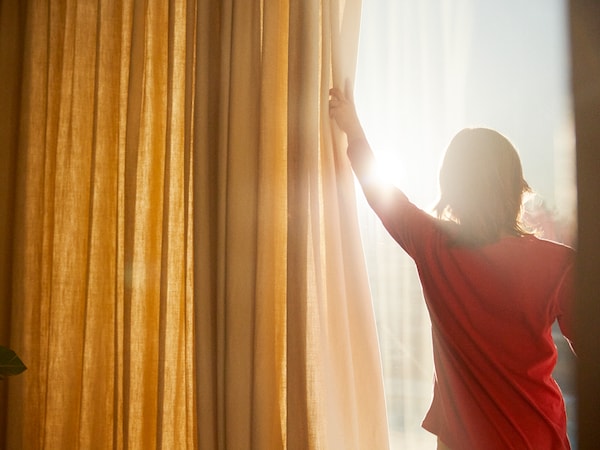 一个孩子在一个红色t恤拉回黄色和白色的窗帘,露出太阳炽热的窗外。