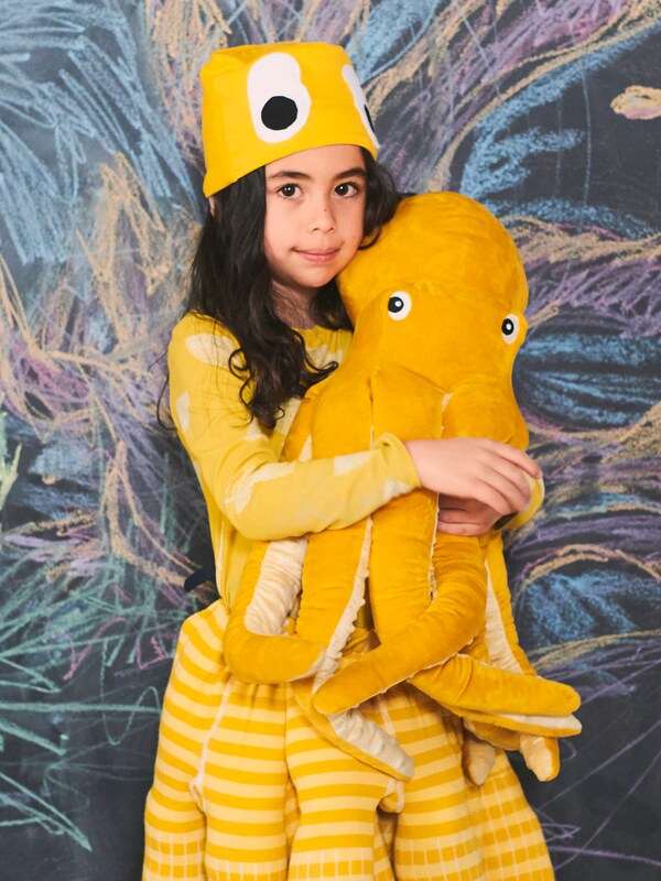 一个女孩穿着黄色衣服拥抱一个黄色BLAVINGAD章鱼毛绒玩具用颜色画在黑板前。