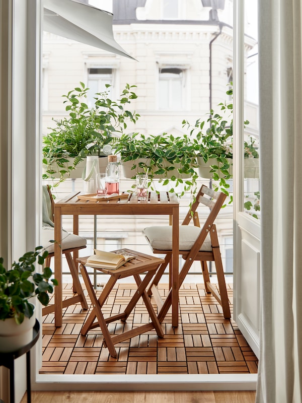 一个light-brown-stained NAMMARO户外表,可折叠凳子和可折叠的椅子在阳台上笼罩在绿叶植物。